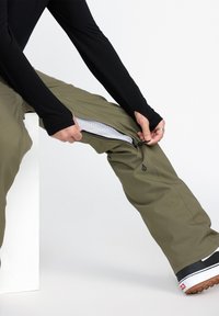 Pantalons vert olive avec des côtés zippés, dotés d'une doublure en mesh. Le modèle porte un haut noir ajusté et des chaussures noires avec des semelles blanches.