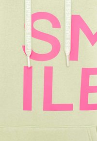 Grüne Kapuzenjacke mit fettem pinken Text, der "SMILE" liest. Schnürsenkel in hellgrün mit "HAPPINESS" in Weiß bedruckt. Weicher Stoff, lässiges Design.