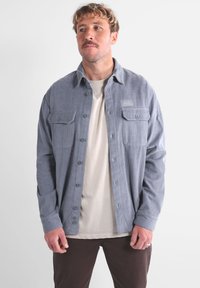 Camisa de pana gris con botones en la parte delantera, dos bolsillos en el pecho y un corte relajado. Llevada sobre una camiseta de color claro.