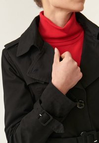 Czarny płaszcz tren dżerowy z podwójnym zapięciem, dużymi klapami i paskiem w talii. Pod spodem widoczny czerwony golf. Gładka tekstura materiału.