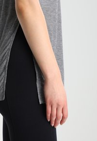 Grijze, gestructureerde top met een losse pasvorm en een zijsplit, gecombineerd met zwarte leggings. Close-up van een hand die op de dij rust.