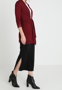 Bordowa bluzka kopertowa z koronkową lamówką, noszona z czarną midi spódnicą z bocznymi rozcięciami oraz czarnymi szpiczastymi botkami za kostkę.