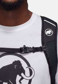 Svart ryggsäkerrem med strukturerad yta, utrustad med en plastklämma och en cirkulär logotyp av en mammut mot en vit t-shirt.