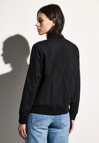Svart bomberjacka med en slät yta, ribbade ärmslut och midja. Modellen har på sig ljusblå jeans och ses från ryggen.