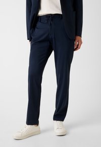 Marineblaue Stretchhose mit Kordelzugbund, zwei Seitentaschen und schmaler Passform, kombiniert mit weißen Sneakern. Glatte Stoffstruktur.