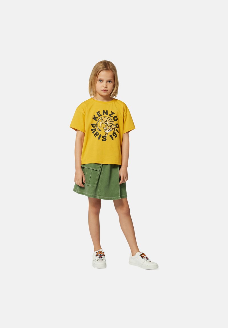 T-shirt en coton jaune avec un graphique de tigre noir et du texte, associé à une jupe en denim verte et des baskets blanches avec des accents brodés.