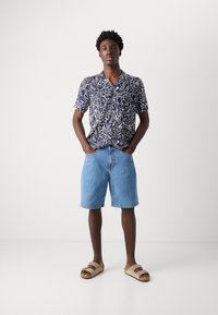 Camisa de manga curta azul-marinho com padrão abstrato branco, combinada com calções de ganga azuis claros e sandálias de dedo cor de tan.