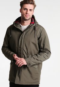 Veste olive avec capuche, dotée d'une doublure à carreaux rouges et noirs, fermeture éclair frontale, cordons de serrage et poches latérales.