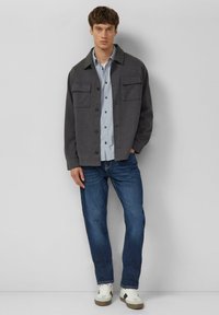 Grijze blousejas met twee borstzakken over een lichtblauw overhemd, gecombineerd met blauwe spijkerbroek en witte sneakers.