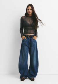 Svart genomskinlig långärmad topp över en mörk bralette, kombinerad med vida blå denimjeans och ett brunt bälte, tillsammans med bruna skor.