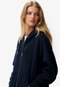 Sudadera con capucha y cremallera en azul marino. Fabricada con un tejido suave y de corte relajado, presenta bolsillos frontales y un logotipo sutil.