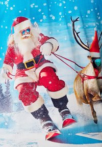 Santa Claus v červeném obleku s bílými kožešinovými doplňky lyžuje na sněhu a vede soba se slunečními brýlemi. V pozadí je zasněžená krajina.