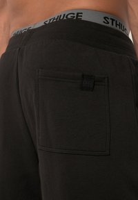 Schwarze Jogginghose mit glatter Textur, ausgestattet mit einer Gesäßtasche und einem grauen Bündchen, auf dem der Schriftzug "STHUGE" gedruckt ist.