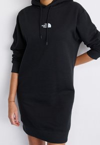 Svart hoodie-klänning med avslappnad passform, tillverkad av mjukt tyg. Har en framstående logo, långa ärmar och ribbade cuffs och nederkant.