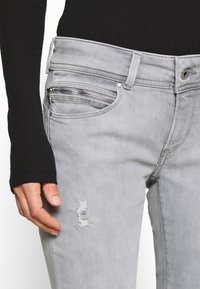 Szare dżinsy z denimu o wąskim kroju. Posiadają małą kieszonkę na zamek oraz przetarcie nad kolanem. Pętle na pasek i zapięcie na guzik z przodu.