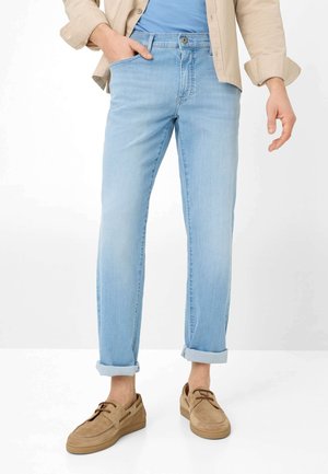 Uomo che indossa jeans azzurri risvoltati, scarpe casual beige, maglietta blu e giacca beige con una mano in tasca.