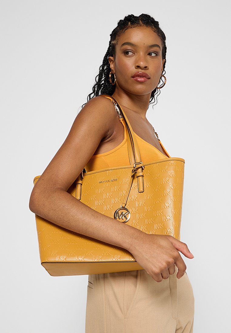 Frau mit geflochtenem Haar, die ein oranges Tanktop und beigefarbene Hose trägt, während sie eine große gelbe Michael Kors Tragetasche über der Schulter trägt.