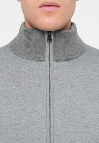 Γκρίζα ζακέτα με φερμουάρ, με χοντρό ριγέ γιακά, που διαθέτει λεία υφή και μπροστινή λεπτομέρεια με φερμουάρ. Απλός, διακριτικός σχεδιασμός.