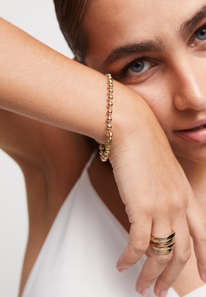 Bracelet en perles dorées sur un poignet, avec un fermoir. Près du bracelet, une bague en or avec des bandes superposées sur un autre doigt. Texture de la peau visible.