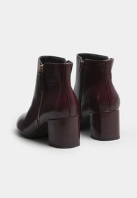 Un par debotines oscuros burdeos con tacones cuadrados y cremalleras laterales, mostrados desde la parte posterior sobre un fondo blanco.