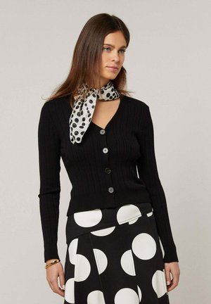 Vrouw die een zwart vest met knopen draagt, een zwarte rok met grote witte stippen en een witte sjaal met zwarte stippen, staand tegen een effen achtergrond.