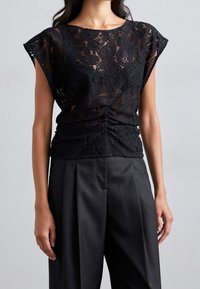 Femme portant un haut en dentelle sans manches noir avec des motifs floraux et un pantalon large plissé noir, sur fond gris uni.