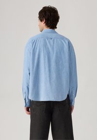 Lyseblå chambray-skjorte med knap-ned krave, lange ærmer og let buet kant; båret med mørke denimbukser.