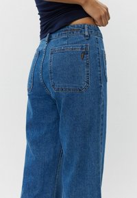 Siniset farkut rentoa istuvuutta, joissa on takataskut ja kirjailtu logo. Kangas on hieman haalistunut.