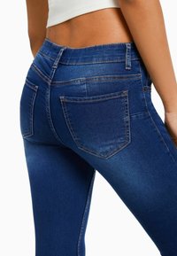 Jeans de mezclilla azul oscuro con un diseño ajustado, que presentan costuras naranjas en contraste y dos bolsillos traseros. Textura suave con un ligero desgaste.