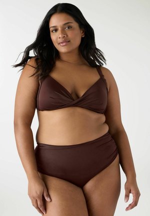 Donna curvy con capelli lunghi e neri che indossa un costume da bagno a due pezzi marrone scuro, in piedi davanti a uno sfondo bianco neutro.