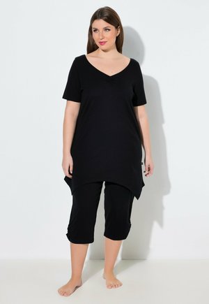 Kvinde står barfodet mod en hvid væg iført en sort V-hals tunika og sorte cropped bukser, kigger let til siden.