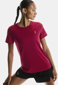 Atletska žena z pletenicami, oblečena v rdečo majico s kratkimi rokavi in črne kratke hlače, pozira v stranskem raztegu pred belo ozadjem.