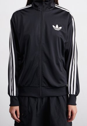 Veste de survêtement noire Adidas avec trois bandes blanches sur les manches et logo trèfle sur le côté gauche de la poitrine, portée zippée par une personne.