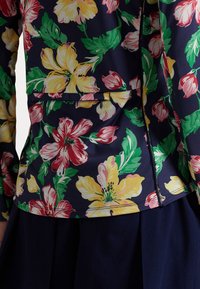 Blusa floral con un fondo marino y grandes flores en tonos rosa, amarillo y verde. Incluye un bolsillo frontal y una tela texturizada.