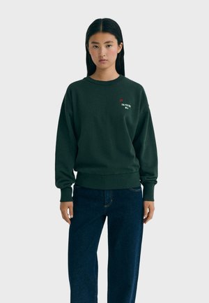 Giovane donna dai capelli neri lisci indossa un felpa verde scuro con un piccolo fiore e testo e jeans blu scuro su uno sfondo uniforme.