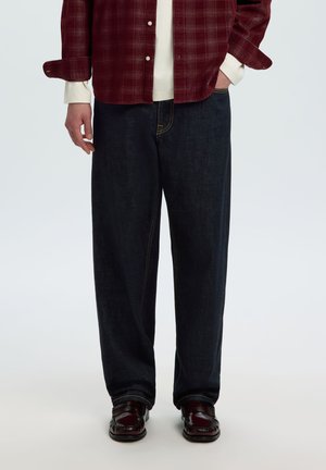 Mand iført mørke jeans med vide ben, burgundy loafers, hvid skjorte og rødternet skjorte med hænderne i lommerne.