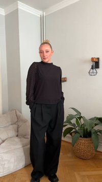 Top nero a maniche lunghe a strati con motivo texturizzato, abbinato a pantaloni neri a vita alta e gamba larga. Scarpe nere solide, ambientazione interna.
