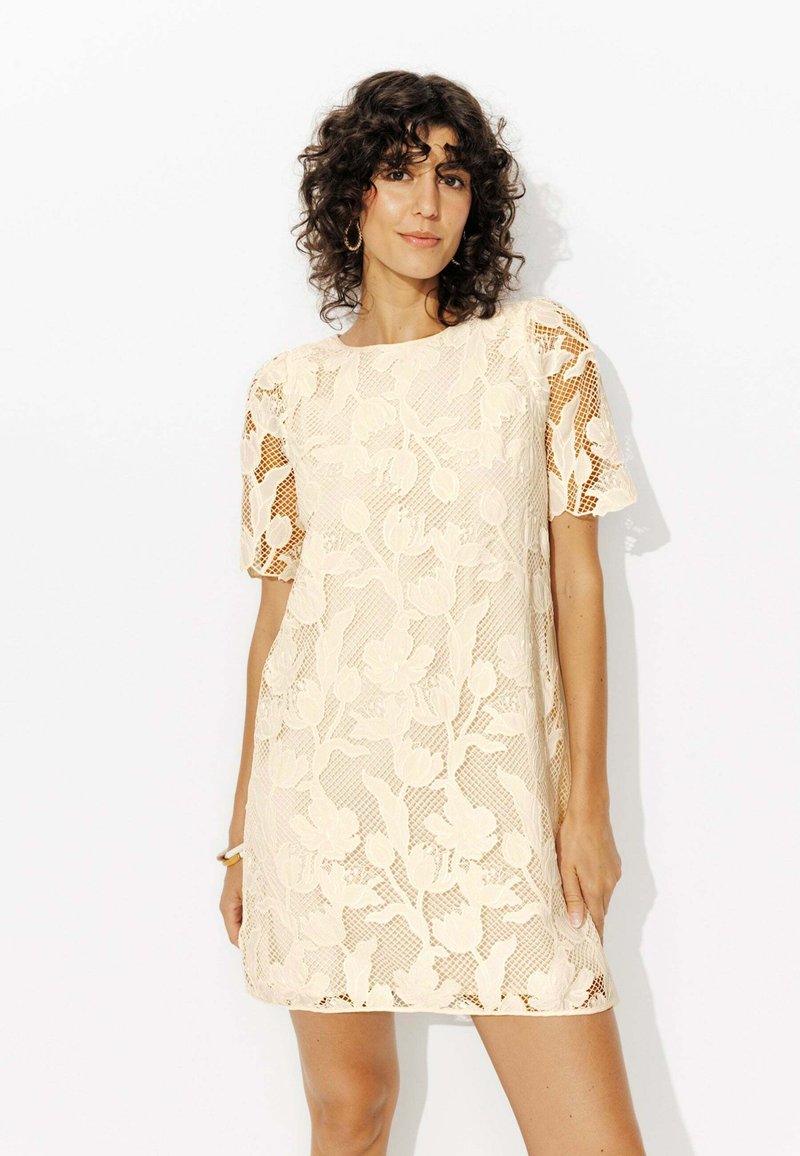Robe en dentelle couleur crème clair avec motif floral, manches courtes, coupe ample et col rond. Tissu texturé avec des accents transparents.