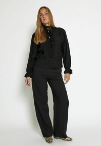 Blusa nera a maniche lunghe con scollo arricciato, abbinata a pantaloni neri a palazzo. L'outfit presenta un tessuto testurizzato e scarpe con stampa animale.