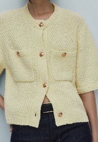 Żółty kardigan na krótki rękaw z dzianiny, z fakturowanym wzorem, dwiema kieszeniami na piersi i brązowymi guzikami. Noszony na ciemnym dżinsie.