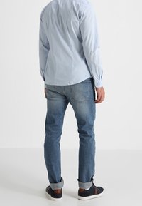 Helles blaues Hemd mit langen Ärmeln, kombiniert mit dunklen Jeans und schwarzen Turnschuhen, die hochgekrempelte Ärmel haben.