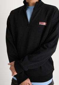 Uomo che indossa un maglione zip-up nero con colletto e polsini a coste, sovrapposto a una camicia azzurro chiaro, con il logo "HUGO" in rosso e bianco sul petto.