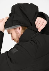 Svart vadderad jacka med justerbar huva. Har en slät yta, lätt glans och en dragkedja vid halsen.