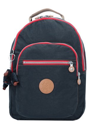 Navy backpack with red accents, zippered pockets, and a furry monkey keychain. Features a circular leather logo patch on the front.