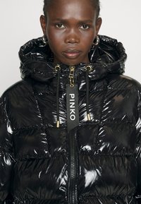 Czarna kurtka puchowa o błyszczącej powierzchni, z kapturem, złotymi detalami i kontrastową metką z logo na suwaku.