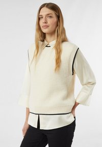 Kremowy dzianinowy sweter bez rękawów z prążkowaną fakturą, z czarnym kontrastowym obszyciem. Nosi się go na białej koszuli z kołnierzykiem i czarnych spodniach.