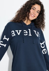 Granatowa bluza z kapturem z długimi rękawami, z białymi napisami na ramionach. Wykonana z miękkiego materiału, zaprojektowana z przednią kieszenią kangurka i kapturem.