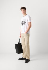 Hvit T-skjorte med svart grafisk design, beige bukser, svart lær totebag og svarte sko med tykk såle. Krøllete hår.