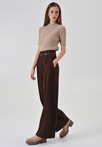 Beiges geripptes Strickoberteil mit kurzen Ärmeln, kombiniert mit hoch taillierten braunen plissierten Hosen. Brauner Gürtel und hellbraune Plateau-Schuhe.