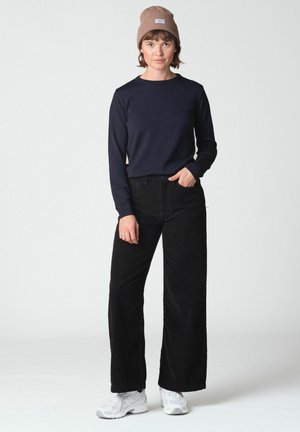 Jeune personne debout avec une main dans la poche, portant un bonnet marron, une chemise à manches longues bleu marine, un pantalon noir à jambes larges et des baskets blanches.